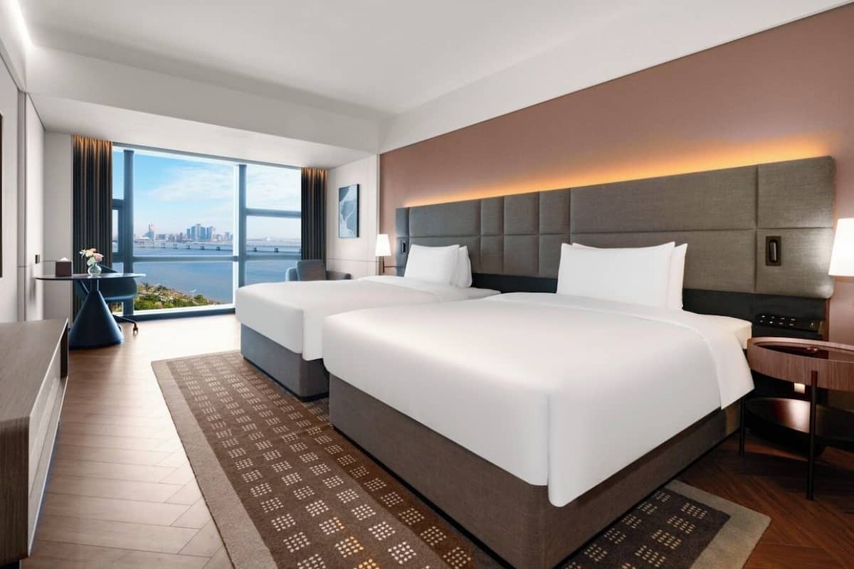 A hotel room with two beds, a large window with a city view, a desk, and a small table with a lamp.