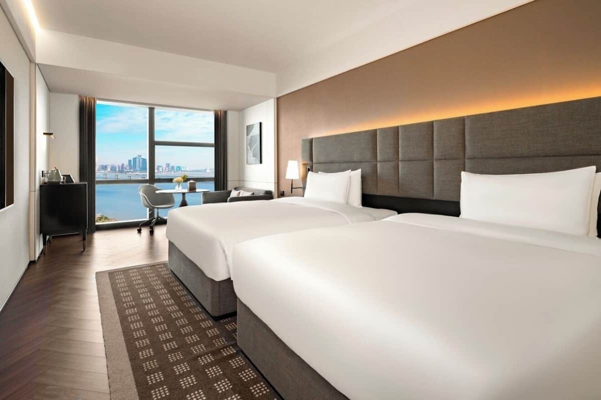 A hotel room with two beds, a desk, a chair, and a view of the city skyline.