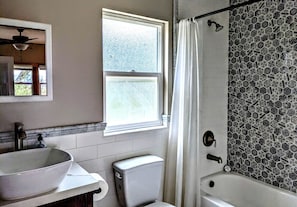Ein Badezimmer mit Waschbecken, Toilette und Badewanne. Über dem Waschbecken befindet sich ein Fenster und ein Duschvorhang.