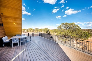 A wooden deck with a table and chairs, a metal railing, and a view of trees and hills.