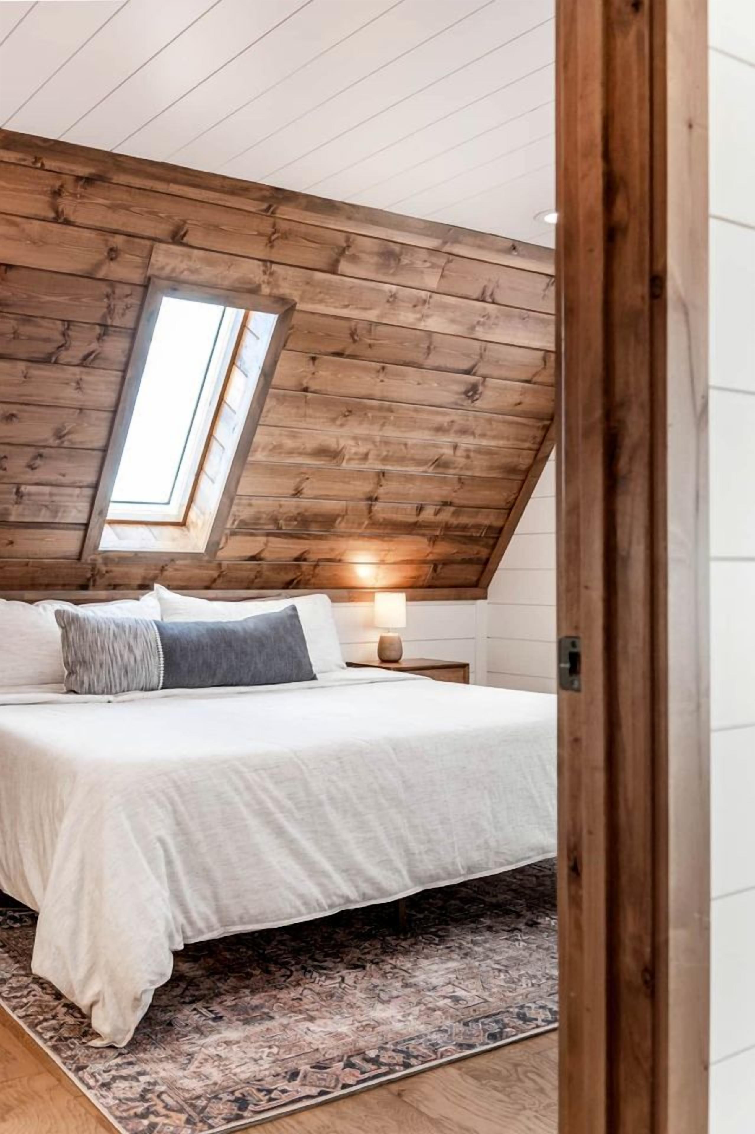 A bedroom with a sloped ceiling, a skylight, a bed with pillows, a bedside table with a lamp, and a patterned rug.