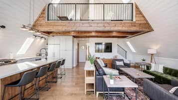 A spacious loft with a high ceiling, a kitchen with a bar area, a living room with a sofa and coffee table, and a staircase leading to an upper level.