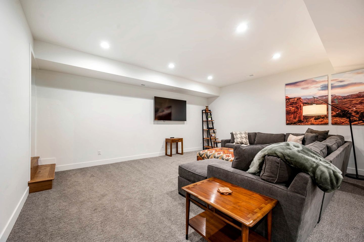 Ein modernes Wohnzimmer mit grauem Sofa, einem Holztisch, einem an der Wand befestigten Fernseher und einem Gemälde einer Wüstenlandschaft.