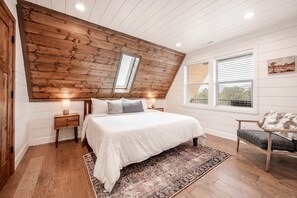 A bedroom with a sloped ceiling, a bed with white bedding, a nightstand, a chair, and a window with blinds.