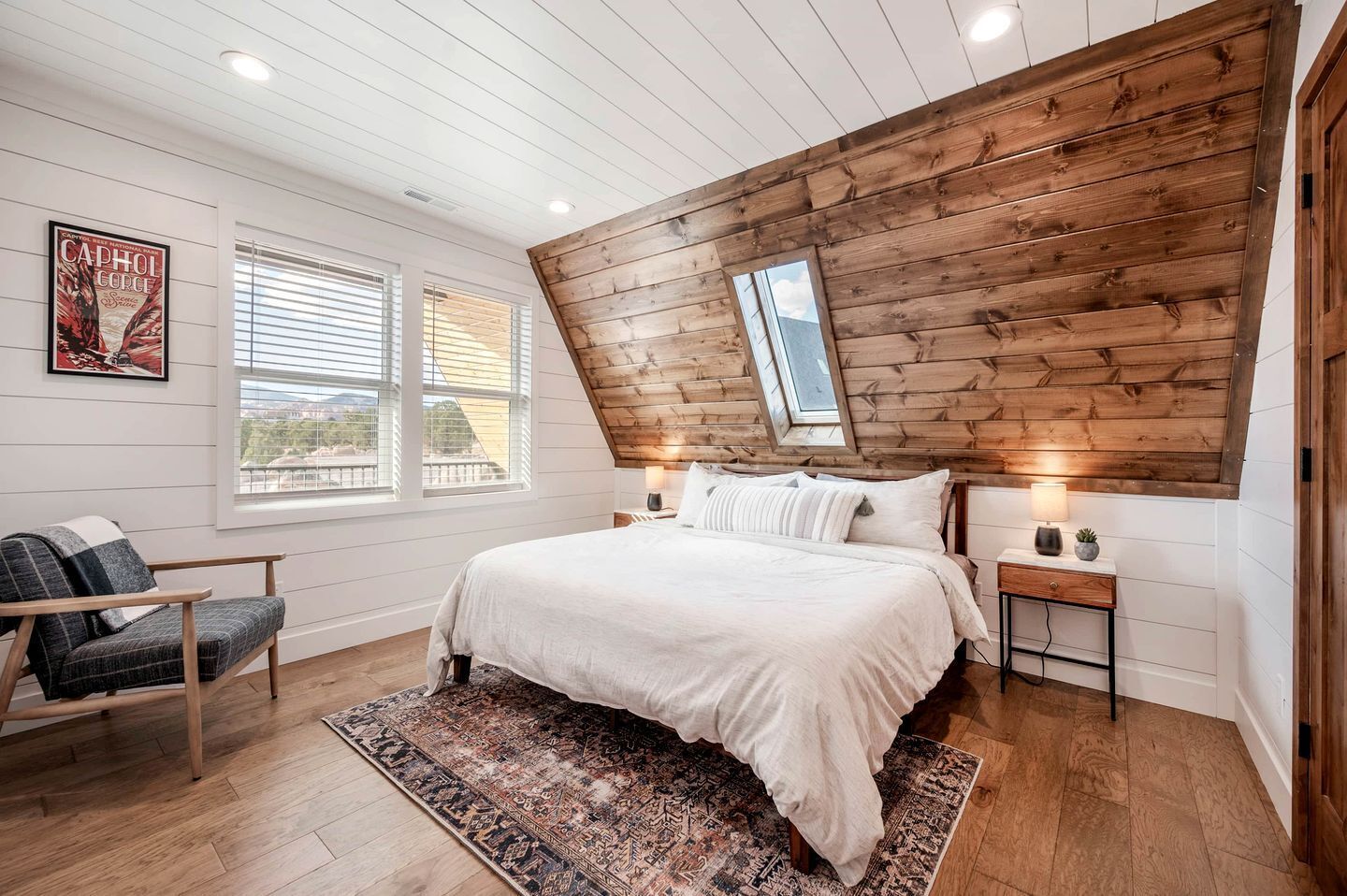 A bedroom with a sloped ceiling, a large bed, a chair, a nightstand, a lamp, and a poster on the wall.