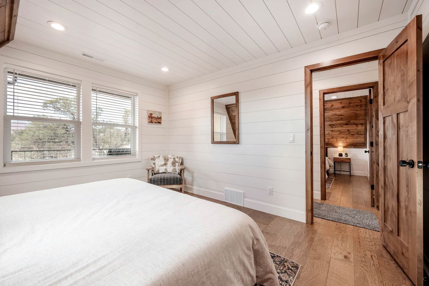 A bedroom with a bed, a chair, a mirror, and a wooden door leading to a bathroom.