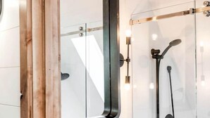 Un bagno moderno con uno specchio a cornice di legno, un lavandino bianco, una piccola pianta in vaso e un box doccia in vetro.