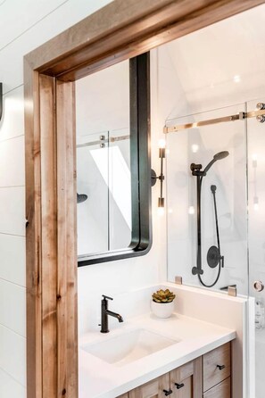 A modern bathroom with a wooden-framed mirror, a white sink, a small potted plant, and a glass shower enclosure. - Experience Pure Glamping on this A-frame Placed in the Middle of the Desert in Wayne County, Utah (Wayne County)