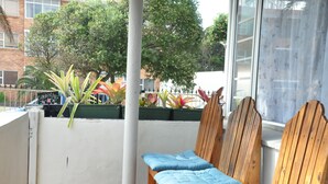 En balkon med træstole og hynder, en hvid væg og udsigt til træer og bygninger.