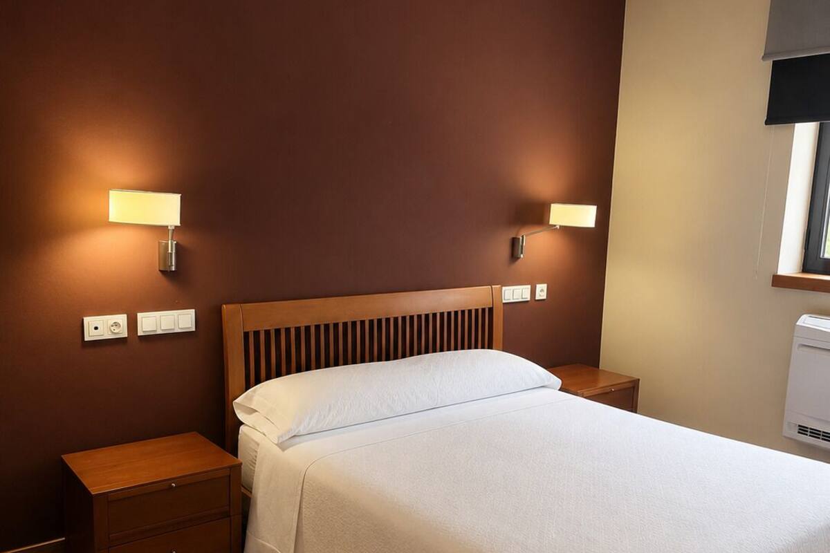 Un dormitorio ordenado con cabecera de madera, dos mesitas de noche, una cama con colcha blanca y lámparas fijadas a la pared.