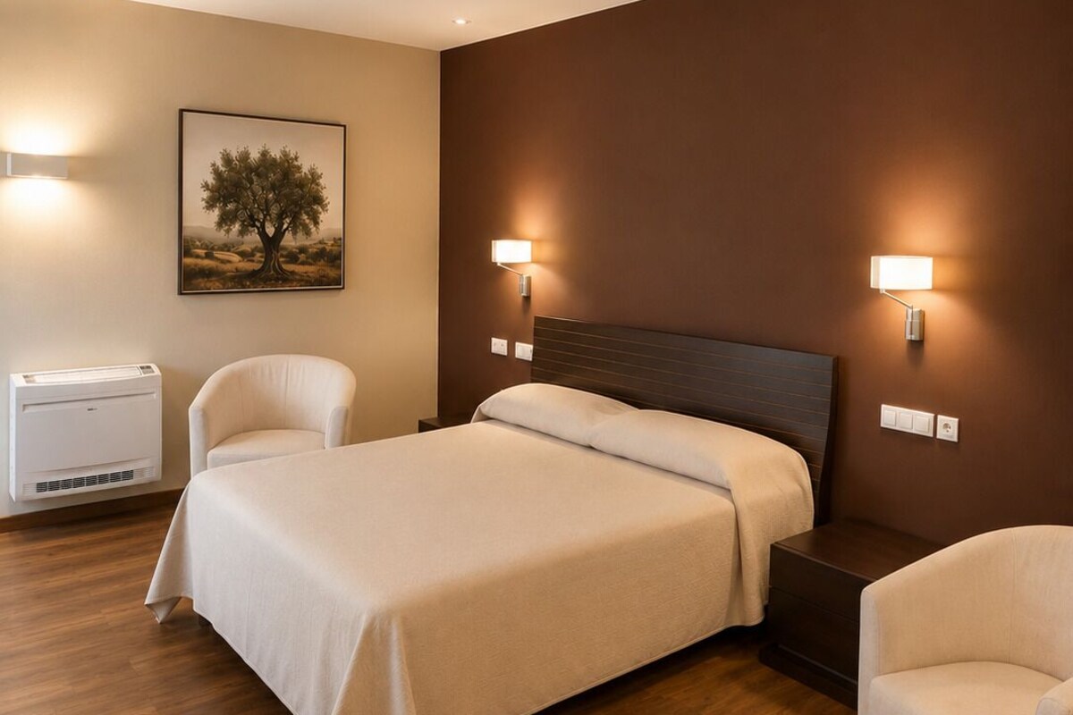 A hotel room with a bed, two chairs, a wall-mounted air conditioner, and a painting.