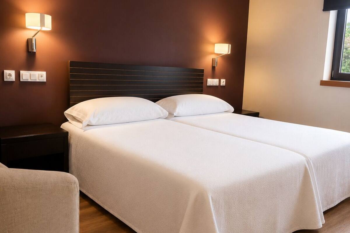 Cama doble con sábanas blancas, cabecera oscura, lámparas de noche y suelo de madera.