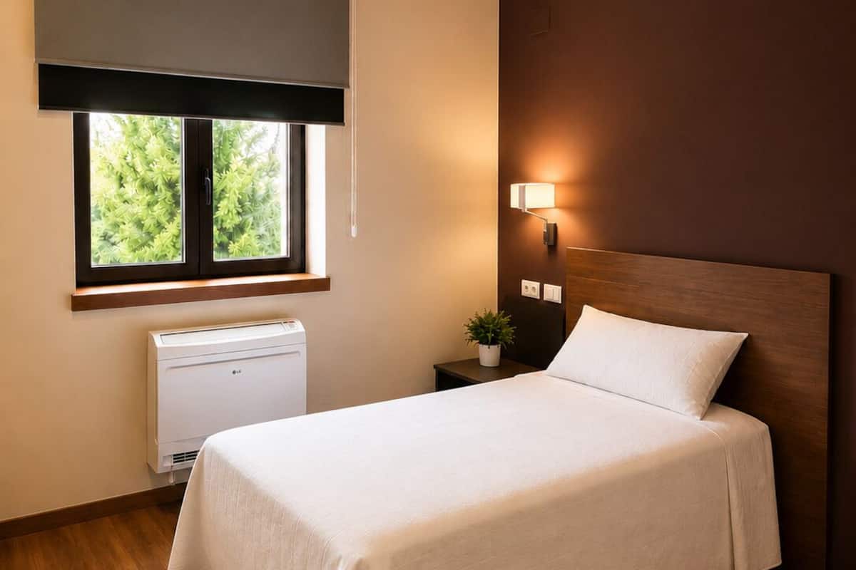 Un dormitorio con cama, ventana con persianas, lámpara de pared y radiador.