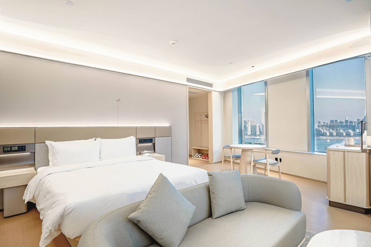 Una habitación de hotel moderna con una cama grande, un sofá, un comedor y vistas a un paisaje urbano.