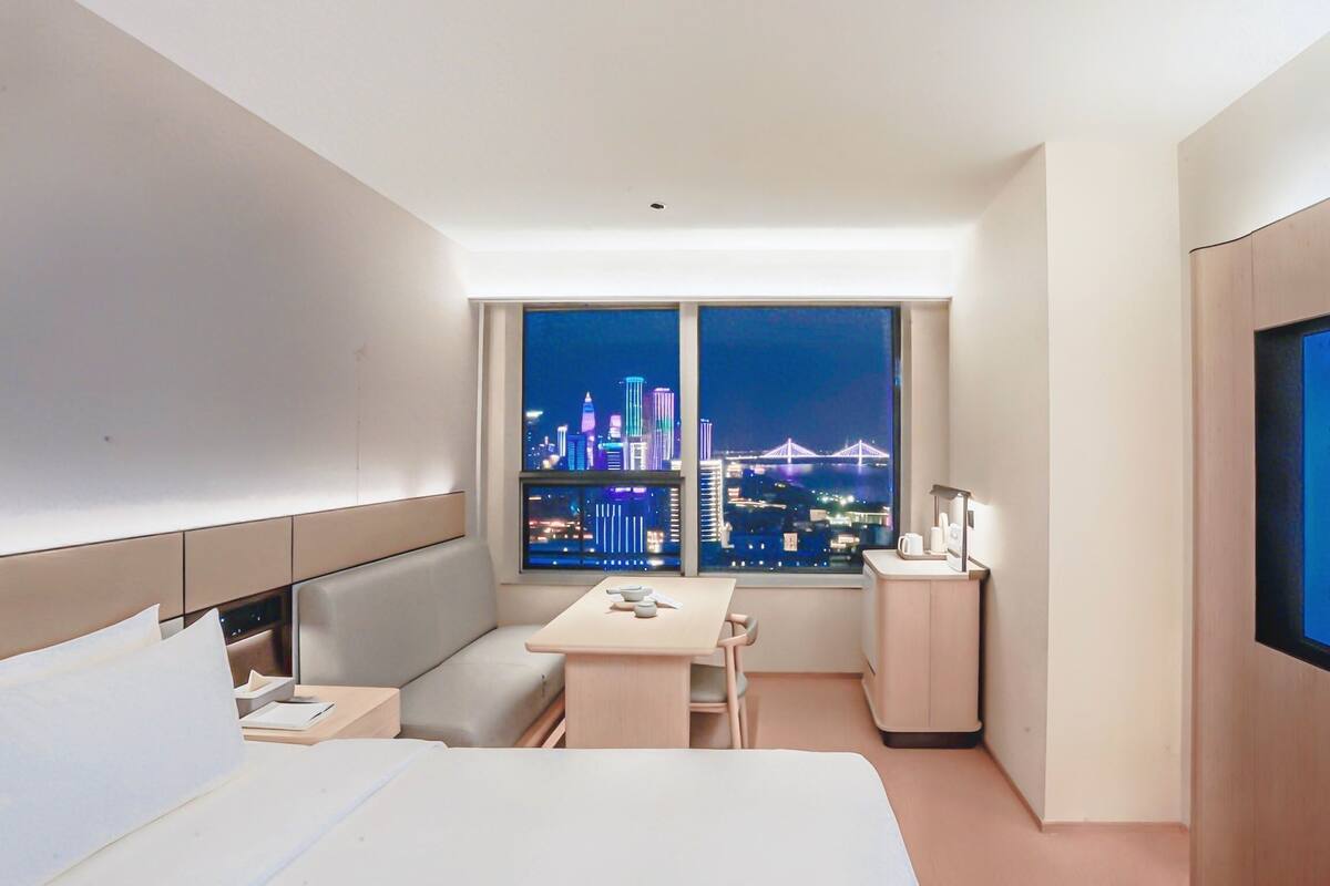 Habitación de hotel con cama, escritorio, silla, una mesita y vistas a un paisaje urbano nocturno.