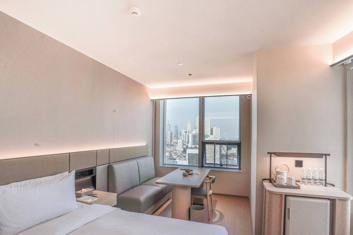 Habitación de hotel con cama, zona de estar, una pequeña mesa, un refrigerador pequeño y vista a un horizonte urbano.