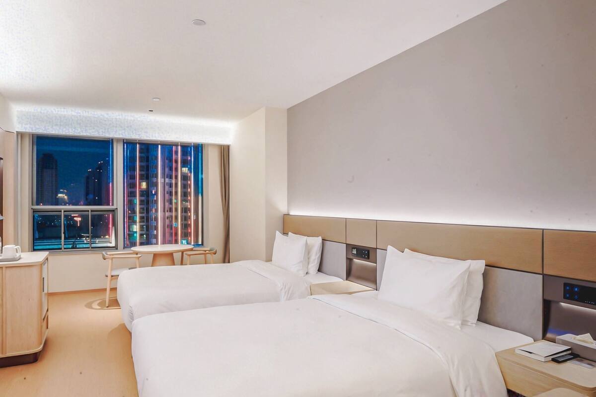 Habitación de hotel con dos camas, un escritorio, una silla y un ventanal con vista a la ciudad.