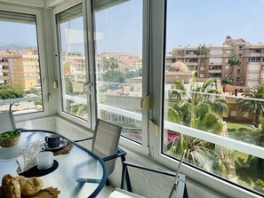 Een balkon met een glazen tafel, stoelen en uitzicht op omliggende gebouwen en palmbomen.