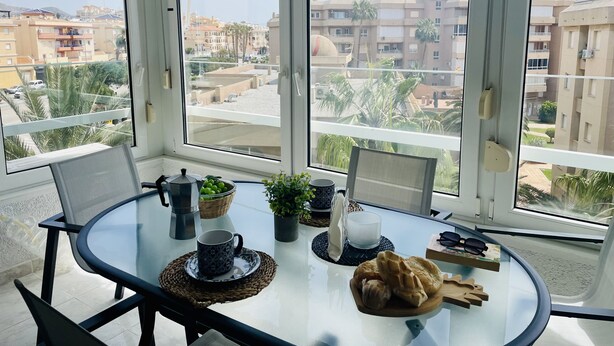 Ein Zimmer mit Glastisch, Stühlen, einer Kaffeemaschine, einer Obstschale und Blick auf Gebäude und Palmen.