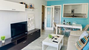 Ein modernes Wohnzimmer mit Flachbildfernseher, Couchtisch, Sofa, Essbereich mit Tisch und Stühlen, Küchenzeile mit Schränken und Geräten sowie Deckenventilator.