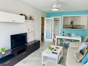 A modern living room with a flat-screen TV, a coffee table, a sofa, a dining area with a table and chairs, a kitchen area with cabinets and appliances, and a ceiling fan. - Goldk Sten by At Home Costa del Sol (Torrox)