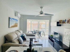 Een moderne woonkamer met een bank, salontafel en een eethoek, grenzend aan een keuken met een televisie en boekenkasten.