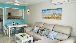 Ein modernes Wohnzimmer mit einer Couch, einem Couchtisch, einem Esstisch und Stühlen. Es gibt einen Deckenventilator, eine Klimaanlage und ein Bild an der Wand.
