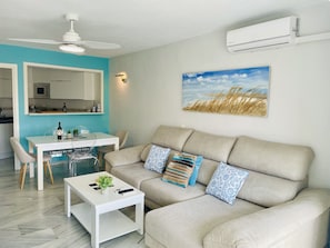 A modern living room with a sofa, coffee table, dining table, and chairs. There is a ceiling fan, an air conditioning unit, and a painting on the wall. - Goldk Sten by At Home Costa del Sol (Torrox)