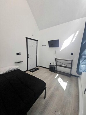 Un dormitorio con cama, una puerta, televisor montado en la pared, una mesita y un cubo de basura.