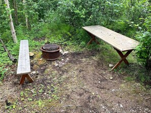A rustic outdoor setting with a wooden bench, a metal fire pit, and a picnic table surrounded by dense greenery.