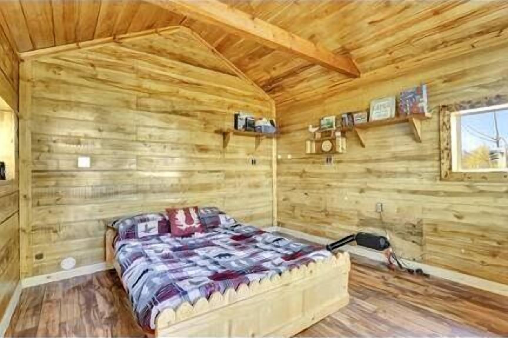 A wooden cabin interior with a bed, shelves with items, and a window.