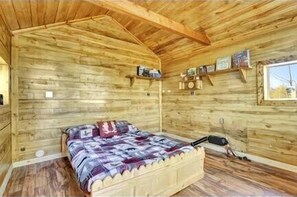 A wooden cabin interior with a bed, shelves with items, and a window.