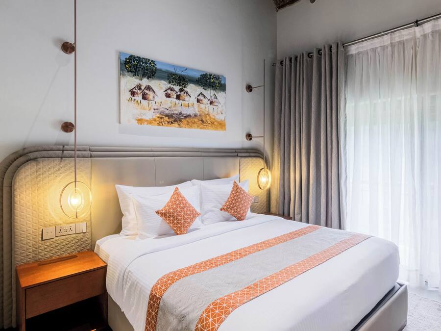 A neatly made bed with a patterned bedspread, a bedside table with a lamp, a wall-mounted painting, and a window with curtains.