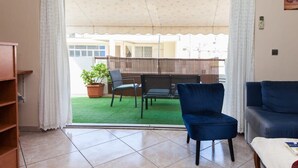 A living room with a blue chair, a sofa, a coffee table, and a view of a patio with a table and chairs. - Cozy & Cute Apartment in Piraeus (Pireas)