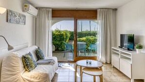 Une pièce comprenant un canapé, une petite table, un téléviseur posé sur un meuble, et une porte-fenêtre coulissante donnant sur un espace extérieur aménagé avec des chaises et de la verdure.