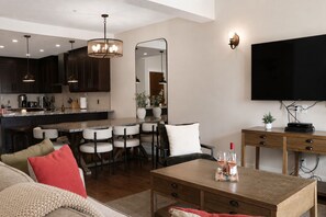 A modern kitchen with dark cabinets, a central island with seating, a dining area with chairs, a flat-screen TV mounted on the wall, and a wooden desk with a plant and wine glasses. - Penthouse with Waterpark | Sleeps 8 | Spacious | Views (Canmore)