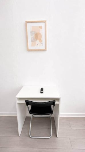 Een wit bureau met een zwarte stoel, een lijst met foto aan de muur en een zwart object op het bureau.