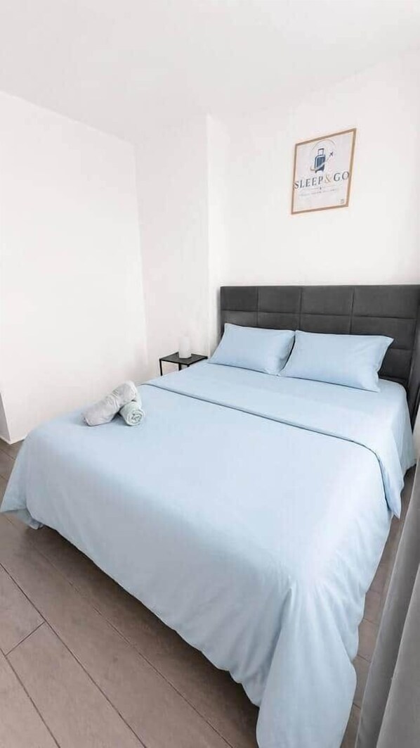 A neatly made bed with a light blue bedspread and pillows, a dark headboard, a small bedside table with a lamp, and a framed sign on the wall. - 5. Bed E5 Center-Station-Airport (Charleroi)