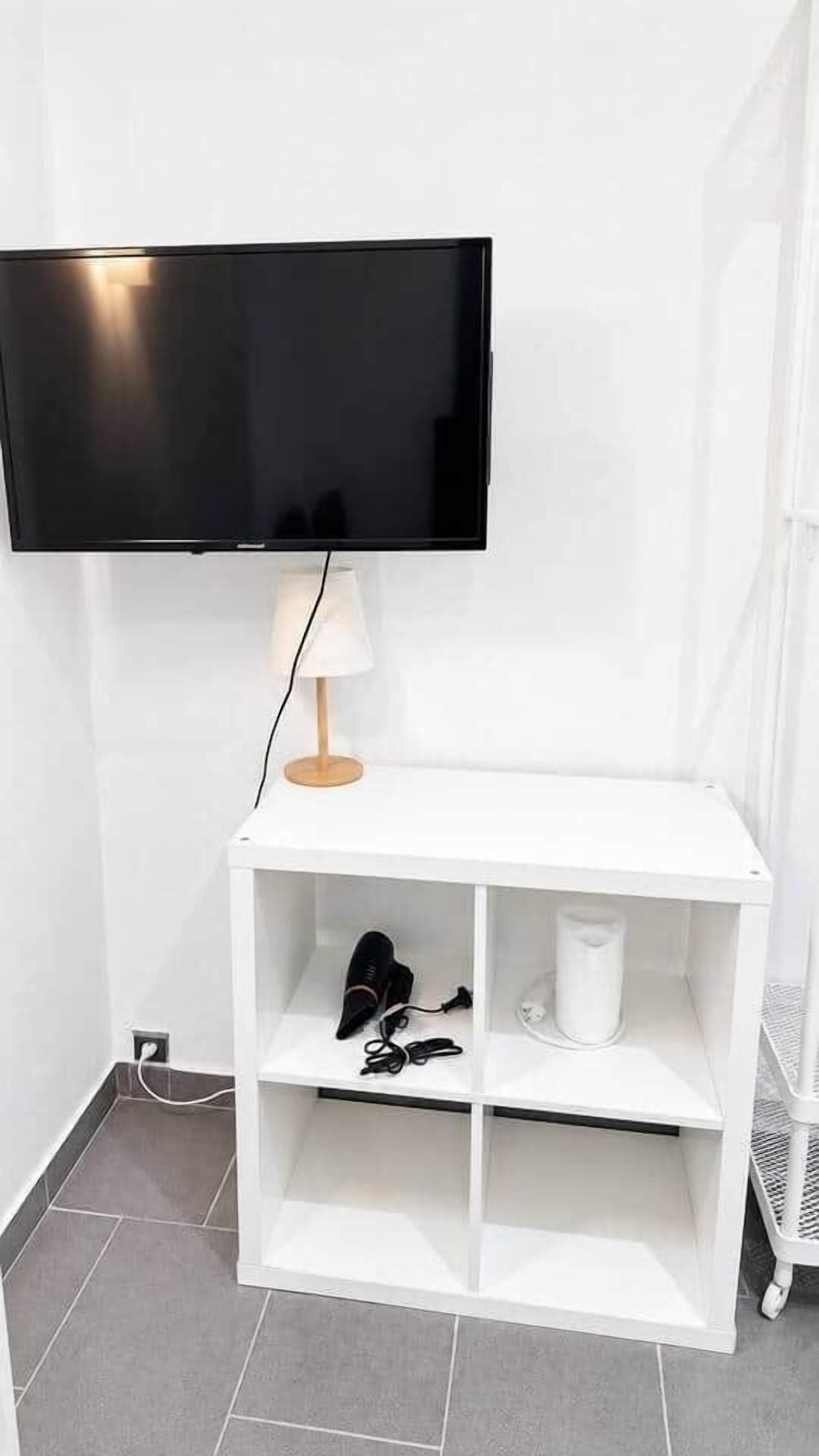 Um móvel branco para TV com uma televisão de tela plana montada acima, uma luminária no móvel e uma prateleira com um secador de cabelo e outros objetos.