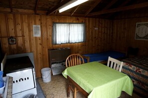 A small cabin with a wooden interior, a table with a green tablecloth, a bed with a patterned blanket, a window with curtains, and a wall clock.