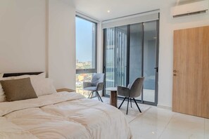 Ein modernes Schlafzimmer mit einem großen Bett, einem kleinen Tisch, zwei Stühlen, einer Schiebetür zum Balkon und einer Holztür.