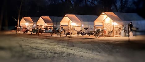 A row of tents with interior lighting, benches, and tables, suggesting a campsite setting.