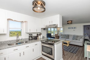 A modern kitchen with white cabinets, a stainless steel sink, and a built-in oven. There is a dining area with a gray sofa and a coffee table. - Sea Breeze enjoy oceanviews and a charming 3-bedroom retreat in Surf City. (Surf City)