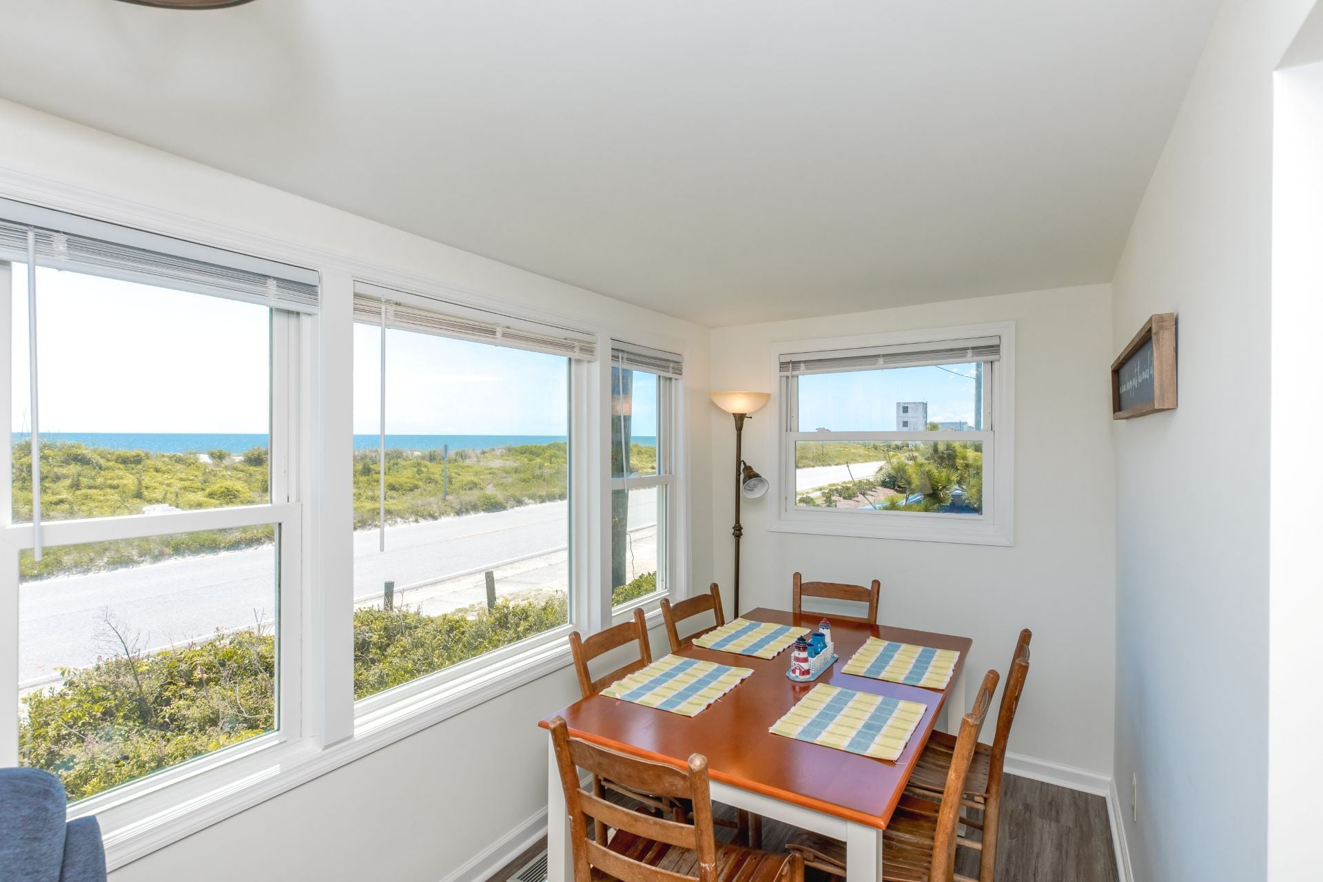 A dining area with a table set for four, large windows with views of the ocean and greenery, and a wall-mounted lamp.
