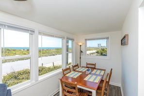 A dining area with a table set for four, large windows with views of the ocean and greenery, and a wall-mounted lamp. - Sea Breeze enjoy oceanviews and a charming 3-bedroom retreat in Surf City. (Surf City)