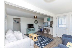 Un salon avec un canapé, une table basse et un téléviseur. Il y a aussi un coin cuisine avec un réfrigérateur, une cuisinière et un panneau indiquant « MAISON DE PLAGE ».