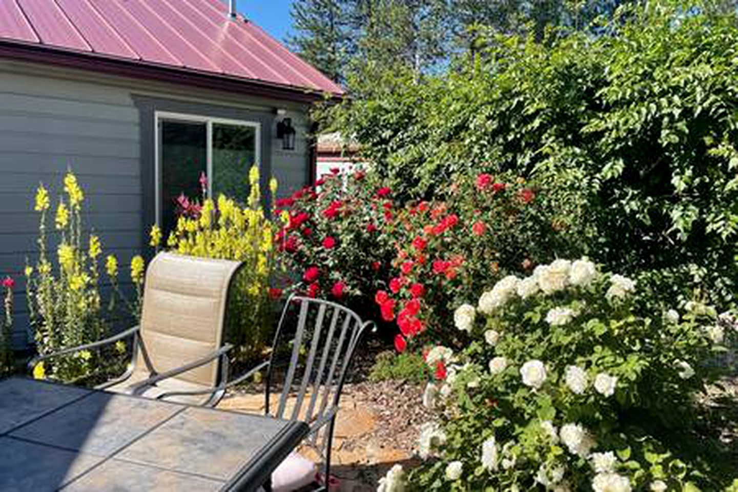 Un patio con una mesa y sillas, rodeado de plantas florecientes y una casa con techo rojo.