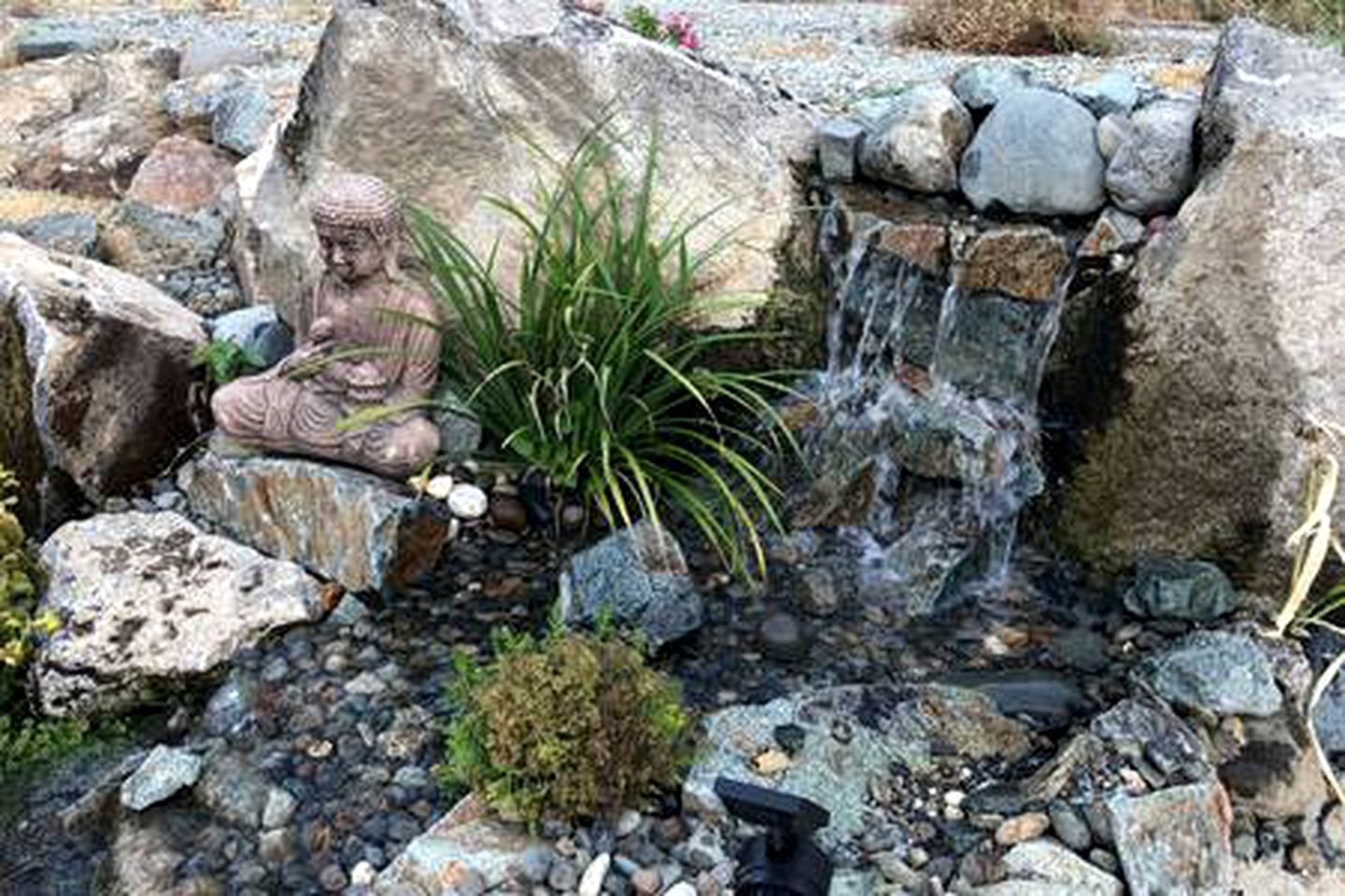 A rock garden with a Buddha statue, a small waterfall, and various plants.