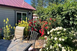 Eine Terrasse mit Tisch und Stühlen, umgeben von blühenden Pflanzen und einem Haus mit rotem Dach.