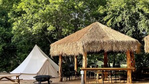 Een camping met een tent, een houten constructie met rieten dak en een picknicktafel.